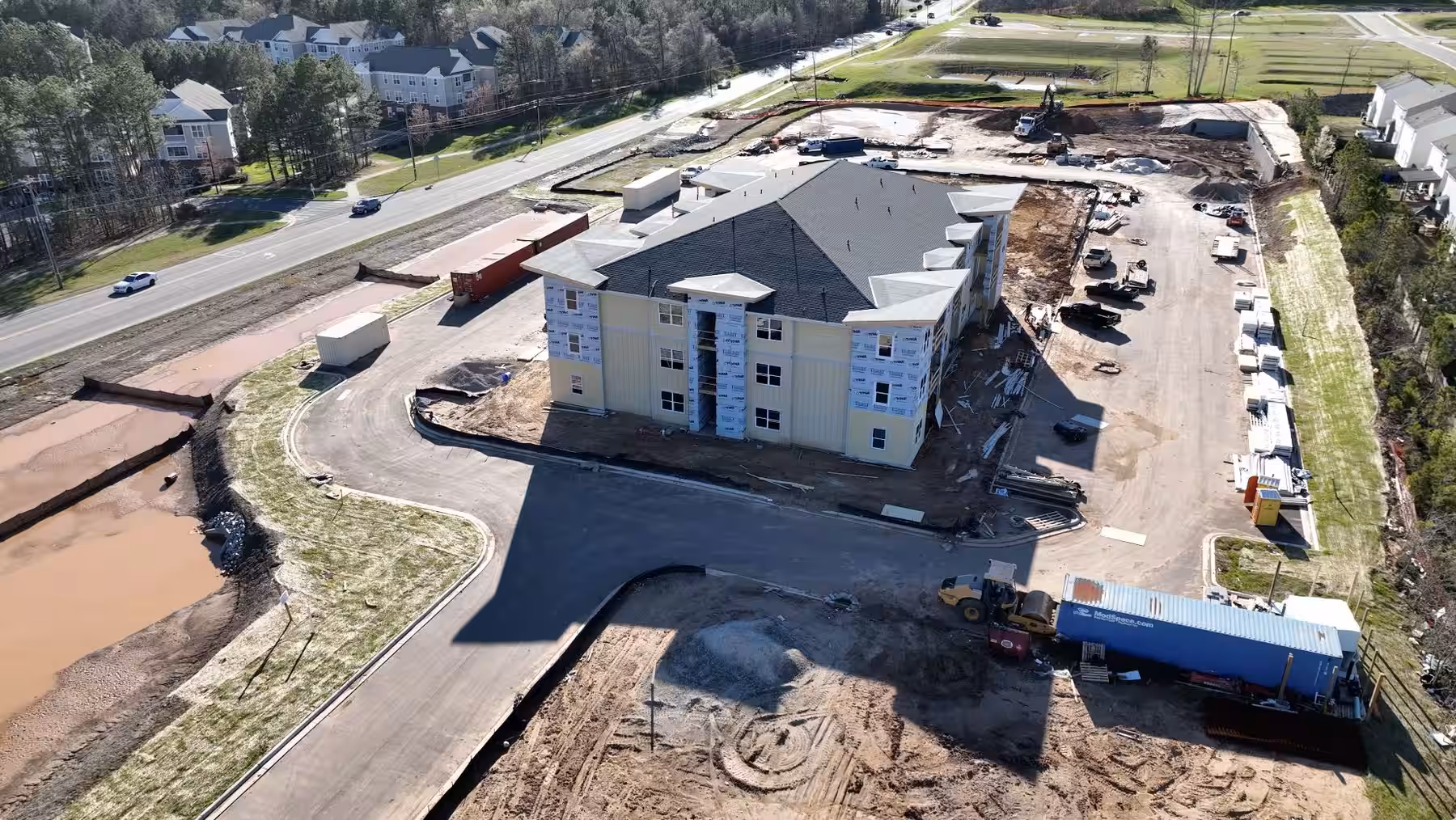 Aerial view of a four-storey apartment building under construction, sheathed and roofed, with graded site-work and a fuel truck on the access road.