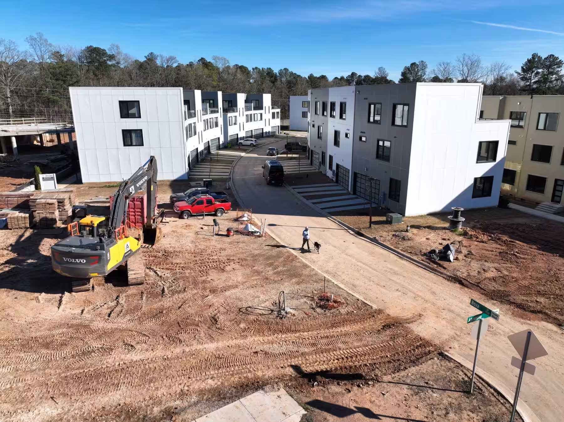 Aerial view of an active residential construction site: framed and sheathed three-storey blocks around a graded interior road, with an excavator working the foreground.