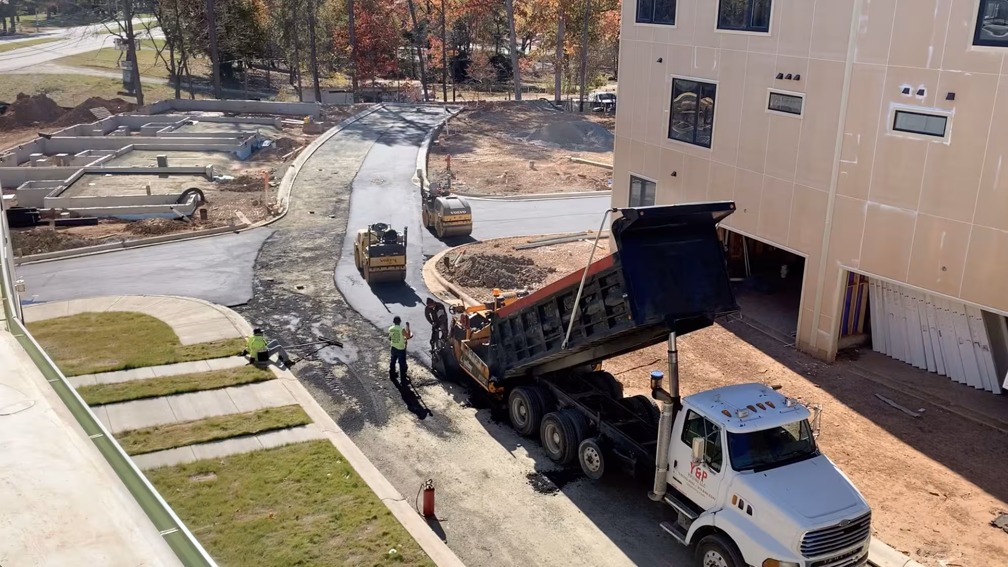 A tri-axle dump truck tipping fill beside a partly clad apartment building, with fresh kerbs, sidewalk and graded roadway running through the site.